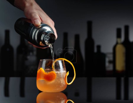 Cocktail Auld Draper auf schwarzem reflektierendem Hintergrund. Im Hintergrund Silhouetten verschiedener Flaschen.