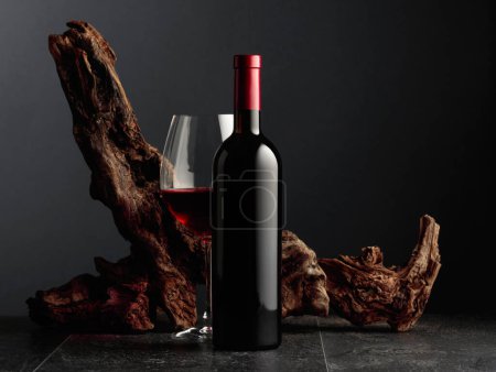 Photo for Bottle and glass of red wine on a black stone table. In the background old weathered snag. Frontal view with copy space. - Royalty Free Image