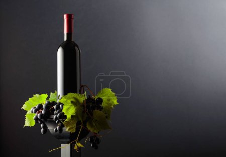 Foto de Botella de rojo en un podio negro. Vino con uvas azules y ramas de vid. Fondo negro con espacio de copia. - Imagen libre de derechos