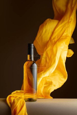 Foto de Botella de vino blanco y aleteos de tela amarilla sobre fondo oscuro. - Imagen libre de derechos