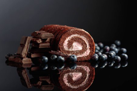 Photo for Chocolate roll cake with blueberries and a broken black chocolate bar on a black reflective background. Copy space. - Royalty Free Image