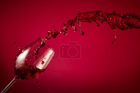 Foto de La copa y el vino tinto salpican sobre un fondo rojo. Copiar espacio. - Imagen libre de derechos