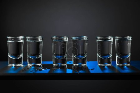 Téléchargez les photos : Coups alcooliques de vodka ou de boisson forte dans de petits verres sur fond noir. Rétro-éclairage bleu. Espace de copie. - en image libre de droit