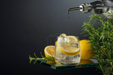 Téléchargez les photos : Cocktail gin tonic avec glace, citron et romarin. Le gin est versé d'une bouteille dans un verre avec des tranches de glace et de citron. Espace de copie. - en image libre de droit