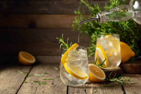 Foto de Cóctel gin tonic con hielo, limón y romero sobre una vieja mesa de madera. La ginebra se vierte de una botella en un vaso con hielo y rodajas de limón. Copiar espacio. - Imagen libre de derechos