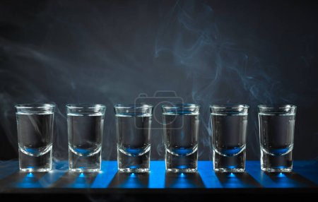 Foto de Las tomas alcohólicas del vodka o la bebida fuerte en los vasos pequeños sobre el fondo negro. Luz de fondo azul. Copiar espacio. - Imagen libre de derechos