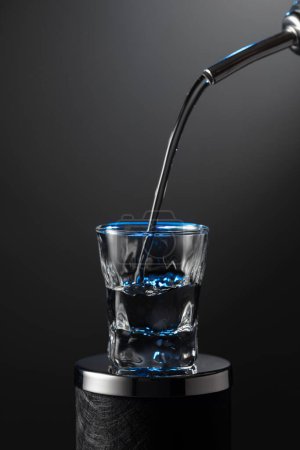 Téléchargez les photos : Vodka verser de la bouteille dans un verre sur un fond sombre. Rétro-éclairage bleu. Espace de copie. - en image libre de droit