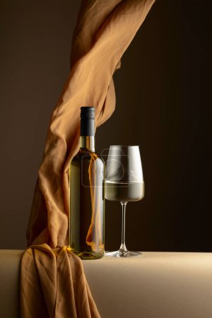 Foto de Botella y copa de vino blanco y aleteos de tela beige sobre fondo oscuro. Copiar espacio. - Imagen libre de derechos