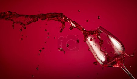 Téléchargez les photos : Verre et vin rouge éclaboussent sur un fond rouge. Espace de copie. - en image libre de droit