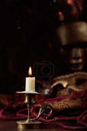 Téléchargez les photos : Bougie allumée dans un vieux chandelier en laiton et masques de carnaval sur une vieille table en bois. - en image libre de droit