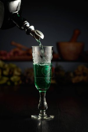 Foto de Licor de hierbas verdes se vierte de una botella vintage en un vaso. En el fondo hierbas secas y utensilios de cocina viejos. - Imagen libre de derechos