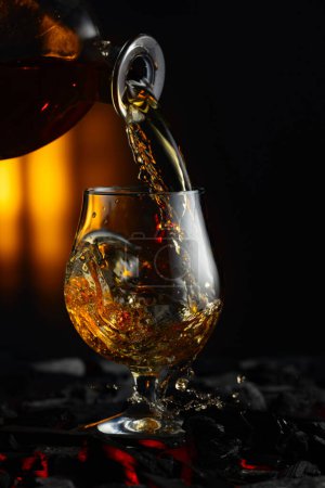 Foto de Se vierte coñac o brandy en un vaso. Snifter en un carbón quemado. Concepto de bebidas alcohólicas duras. - Imagen libre de derechos