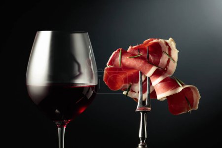 Foto de Copa de vino tinto y jamón en rodajas con romero sobre un tenedor, fondo negro. - Imagen libre de derechos