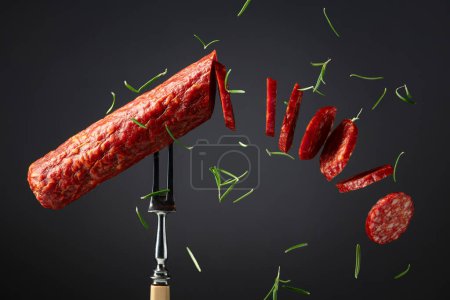 Téléchargez les photos : Saucisse de salami fumée traditionnelle sur une fourchette saupoudrée de romarin. Tranches de saucisse en mouvement. - en image libre de droit
