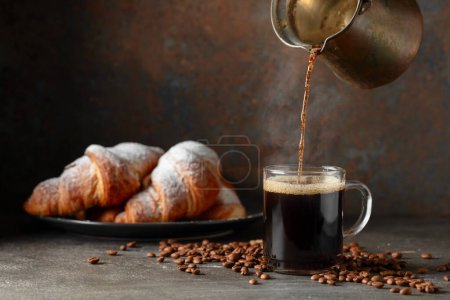 Foto de El café se vierte en una taza. Croissants y café negro en una mesa de la cocina. Concéntrate en un primer plano. - Imagen libre de derechos