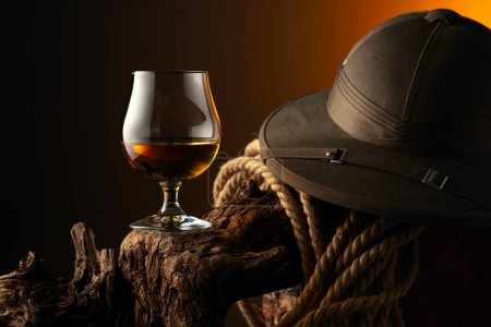 Photo for Snifter of brandy, cork tropical colonial helmet and rope on a old wooden snag. Glass with whiskey, cognac or brandy on dark background. - Royalty Free Image