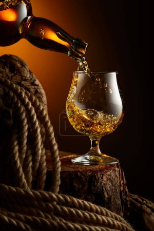 Téléchargez les photos : Le brandy est versé d'une bouteille dans un verre. Verre avec whisky, cognac ou brandy sur une vieille souche. - en image libre de droit