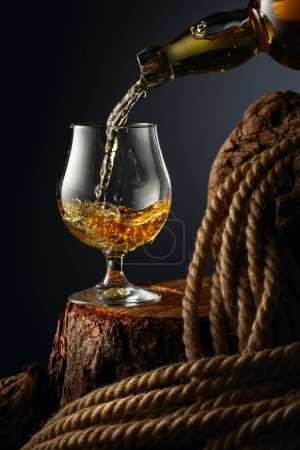 Photo for Brandy is poured from a bottle into a glass. Glass with whiskey, cognac or brandy on an old stump. - Royalty Free Image