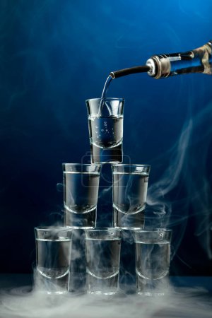 Téléchargez les photos : La vodka est versée d'une bouteille dans un verre en fumée sur fond bleu foncé. Concept de boissons club. - en image libre de droit