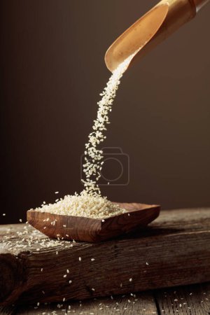 Photo for Grains of white sesame are poured with a wooden spoon in the wooden dish. Copy space. - Royalty Free Image