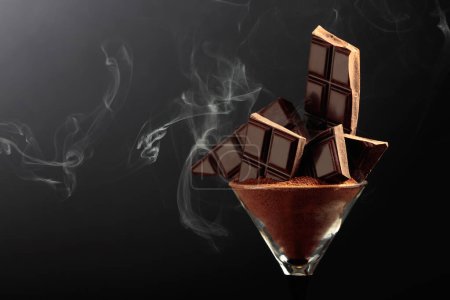 Téléchargez les photos : Barre de chocolat noir cassée en verre avec poudre de cacao. Chocolat sur fond noir avec espace de copie. - en image libre de droit
