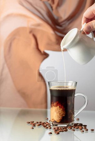 Foto de Café con crema sobre una mesa de cristal. La mano de un hombre derrama crema en un vaso de café. En el fondo ondeando cortina beige. - Imagen libre de derechos