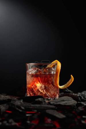 Foto de Cóctel a la antigua con hielo y piel de naranja. Un vaso con un cóctel sobre carbón quemado. Copiar espacio. - Imagen libre de derechos