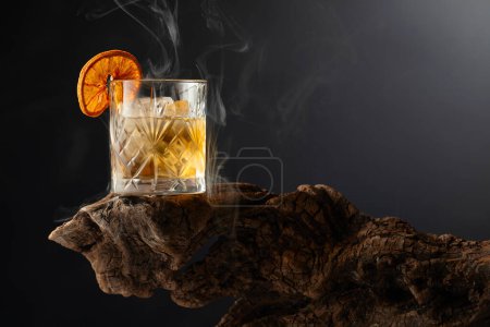 Foto de Cóctel de ron ahumado a la antigua usanza con hielo y rebanada de naranja seca en un viejo enganche de madera. Copiar espacio. - Imagen libre de derechos