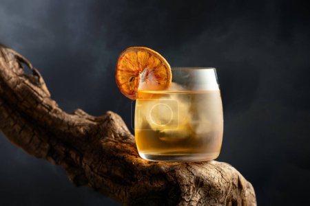 Foto de Cóctel a la antigua con hielo y rebanada de naranja seca en un viejo enganche de madera. Bebida fría sobre un fondo de cielo nublado. - Imagen libre de derechos
