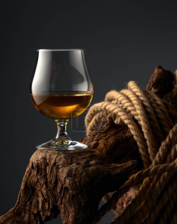 Téléchargez les photos : Brandy snifter et corde sur un vieux chevalet en bois. Verre avec whisky, cognac ou brandy sur fond sombre. - en image libre de droit