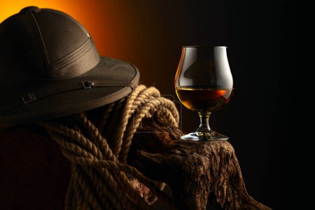 Photo for Snifter of brandy, cork tropical colonial helmet and rope on a old wooden snag. Glass with whiskey, cognac or brandy on dark background. - Royalty Free Image