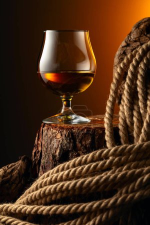 Téléchargez les photos : Un snifter de brandy et une corde sur une vieille tige. Verre avec whisky, cognac ou brandy sur fond sombre. - en image libre de droit