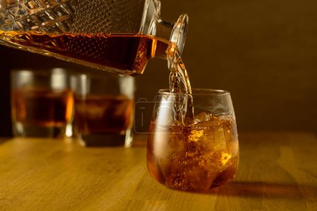 Foto de El whisky se vierte en un vaso embalsado con hielo natural. Un vaso de whisky en una mesa de roble. - Imagen libre de derechos