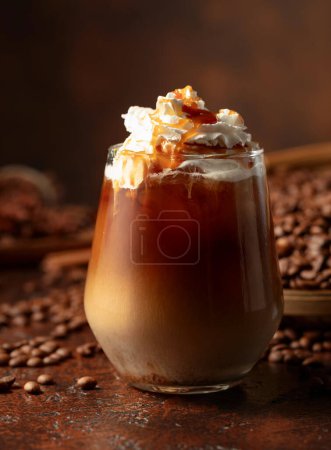Foto de Latte de caramelo helado cubierto con crema batida y salsa de caramelo. Refrescante y dulce bebida de café en una mesa marrón. - Imagen libre de derechos