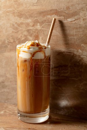 Téléchargez les photos : Latte au caramel glacé garnie de crème fouettée et de sauce caramel, boisson rafraîchissante et sucrée sur une table en céramique beige. - en image libre de droit