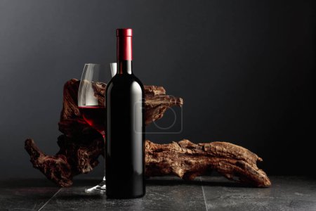 Photo for Bottle and glass of red wine on a black stone table. In the background old weathered snag. Frontal view with copy space. - Royalty Free Image