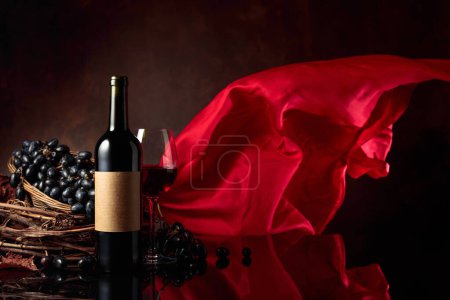 Téléchargez les photos : Verre et bouteille de vin rouge avec raisins sur fond noir réfléchissant. Sur une vieille étiquette vide. Rideau en satin rouge flotte dans le vent. Espace de copie. - en image libre de droit