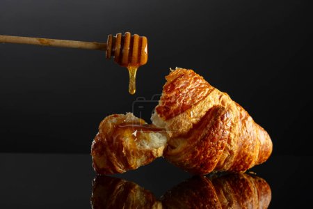Téléchargez les photos : Croissant frais cuit au four avec du miel sur un fond réfléchissant noir. Espace de copie. - en image libre de droit