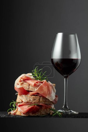 Foto de Ciabatta con jamón, romero y copa de vino tinto sobre fondo negro. Snack italiano tradicional. - Imagen libre de derechos