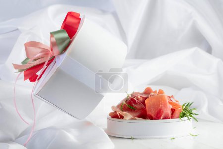 Photo for Prosciutto with rosemary in gift box on a white wooden table. Concept of traditional Italian kitchen. - Royalty Free Image