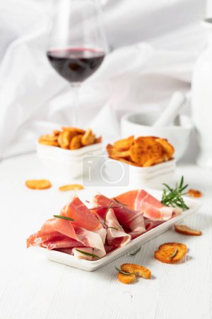 Téléchargez les photos : Du prosciutto, des bruschetta craquelins épicés et un verre de vin rouge sur une table en bois blanc. Cuisine italienne traditionnelle. - en image libre de droit
