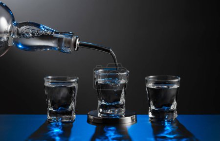 Foto de Vodka que vierte de la botella en un vaso sobre un fondo oscuro. Luz de fondo azul. Copiar espacio. - Imagen libre de derechos