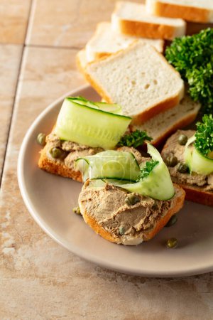 Photo for Toasts with pate on a beige plate. Open sandwiches with pate, fresh cucumber, capers, and parsley. - Royalty Free Image