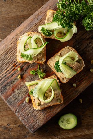 Photo for Toasts with pate on a old cutting board. Open sandwiches with pate, fresh cucumber, capers, and parsley. - Royalty Free Image