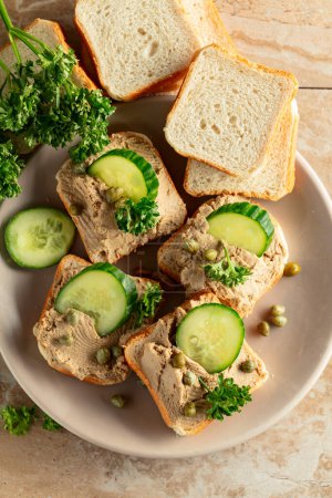 Photo for Toasts with pate on a beige plate. Open sandwiches with pate, fresh cucumber, capers, and parsley. Top view. - Royalty Free Image