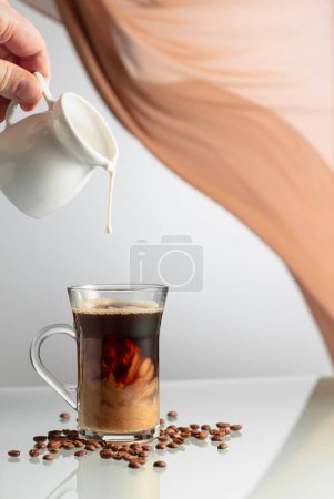 Téléchargez les photos : Café à la crème sur une table en verre. Une main d'homme verse de la crème dans un verre de café. En arrière-plan ondulant rideau beige. - en image libre de droit