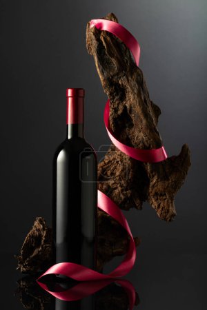 Photo for Bottle of red wine on a black reflective background. In the background old weathered snag with red satin ribbon. Concept of expensive wine. - Royalty Free Image