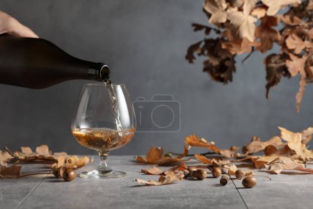 Téléchargez les photos : Le brandy est versé d'une bouteille dans un verre. Snifter de cognac sur une table en pierre aux feuilles de chêne séchées. - en image libre de droit