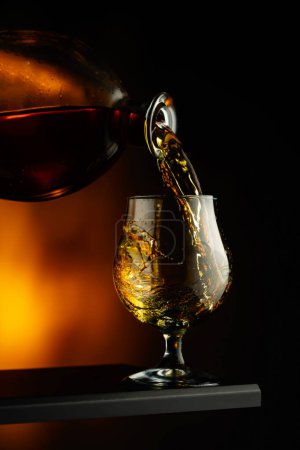 Téléchargez les photos : Cognac ou brandy versé dans un verre. Brandy snifter sur un fond sombre. - en image libre de droit