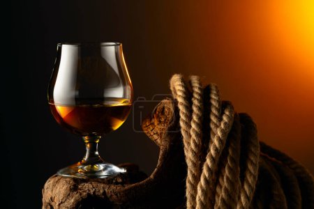 Foto de Brandy francotirador y cuerda en un viejo enganche de madera. Vaso con whisky, coñac o brandy sobre fondo oscuro. - Imagen libre de derechos